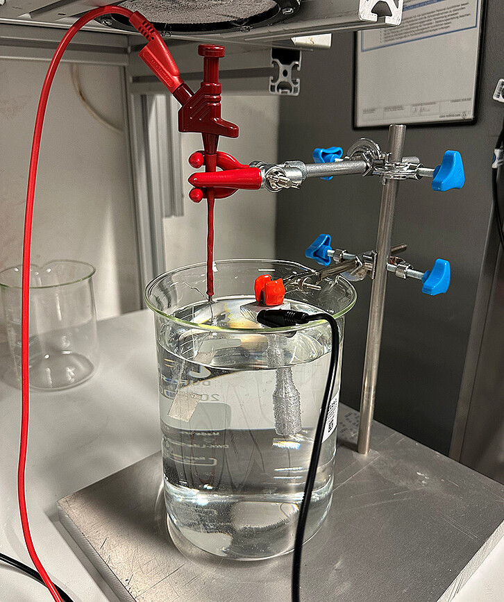 In the laboratory of the Institute for Applied Materials, material tests are carried out on samples loaded with hydrogen by electrolysis. The test setup with liquid bath, sample holder, and power supply is shown. The aim is to identify materials that lose as little fatigue strength as possible under the influence of hydrogen. The tests also investigate which coatings or alloys further reduce the harmful influence of hydrogen. (Image: KIT)
