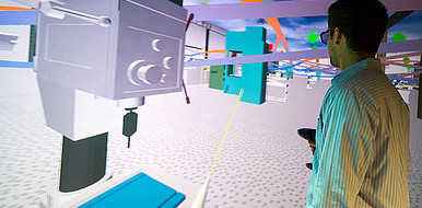 Beispiel: Ein Mensch steht in einer CAVE, ein spezieller Projektionsraum, und navigiert mit einem Controller durch eine virtuelle Fabrik. (Bild: Daniel Schönen / Stadtmarketing Karlsruhe GmbH)