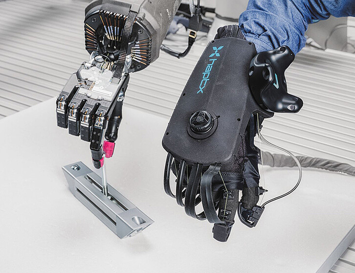Eine Roboterhand (links) lernt per Nachahmung: Mithilfe eines VR-Handschuhs (HaptX, rechts) werden Handlungen von Menschen vorgeführt und vom Roboter synchron nachempfunden. Bereits nach 10 bis 20 Demonstrationen kann der Cobot selbstständig agieren und Aufgaben ausführen. Im Bild montiert der Roboter teleoperativ eine Schraube. (Bild: Amadeus Bramsiepe / KIT)