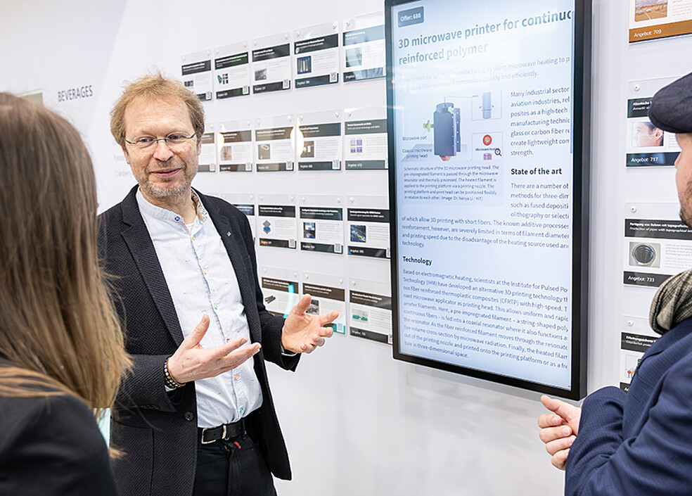 Ein Innovationsmanager des KIT erläutert am Messestand eine Technologieentwicklung auf einem Display, umgeben von Infotafeln mit weiteren Technologieangeboten. Die Technologiewand ist Treffpunkt für Gespräche. (Bild: KIT)